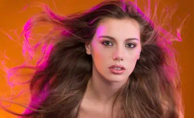 A person with long, flowing hair and a neutral expression is illuminated by pink and orange lighting.