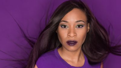 A woman with long dark hair and purple lipstick poses against a purple background, wearing a purple top.