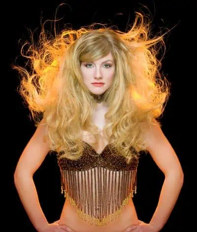 A woman with voluminous blond hair wears a fringed top, posing against a black background with her hands on her hips.