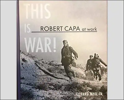 The cover of "This is War!" by Richard Whelan showcases a black-and-white photo of soldiers charging into battle, highlighted by the bold text "Robert Capa at work," celebrating the legendary war photographer. The author's name appears prominently at the bottom.