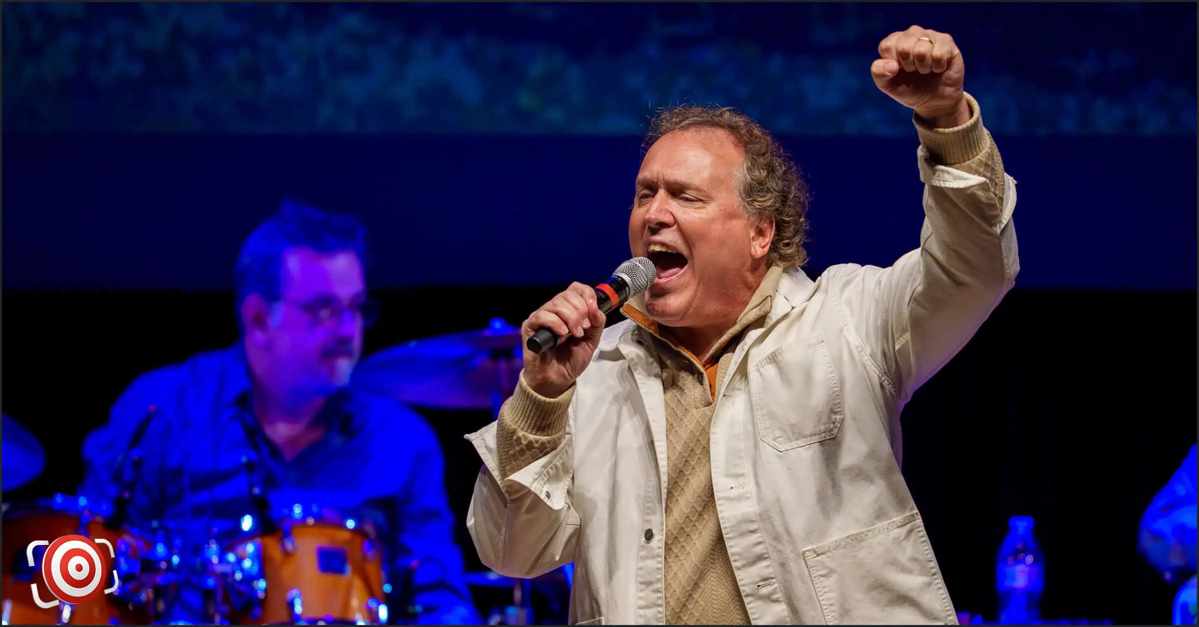 A man in a white jacket, reminiscent of Kenny Rogers, passionately sings into a microphone on stage with the band’s drummer creating an electrifying backdrop.
