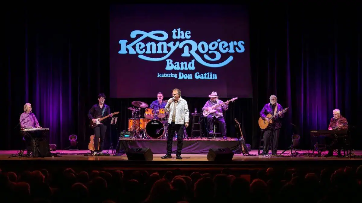 The Kenny Rogers Band performs on stage, featuring Don Gatlin. Members play various instruments under a purple-lit backdrop with the band name displayed above.