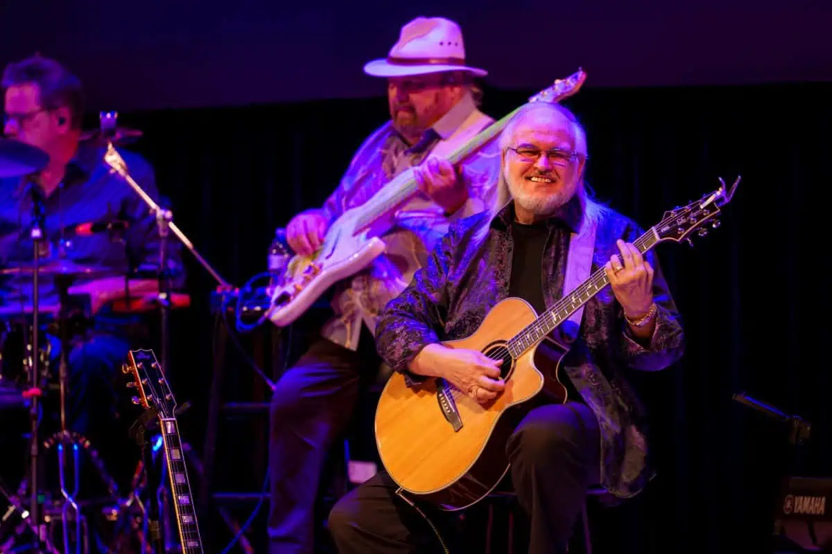 A seated guitarist smiles while playing an acoustic guitar on stage. Behind him, a bassist and drummer perform under bright stage lighting.
