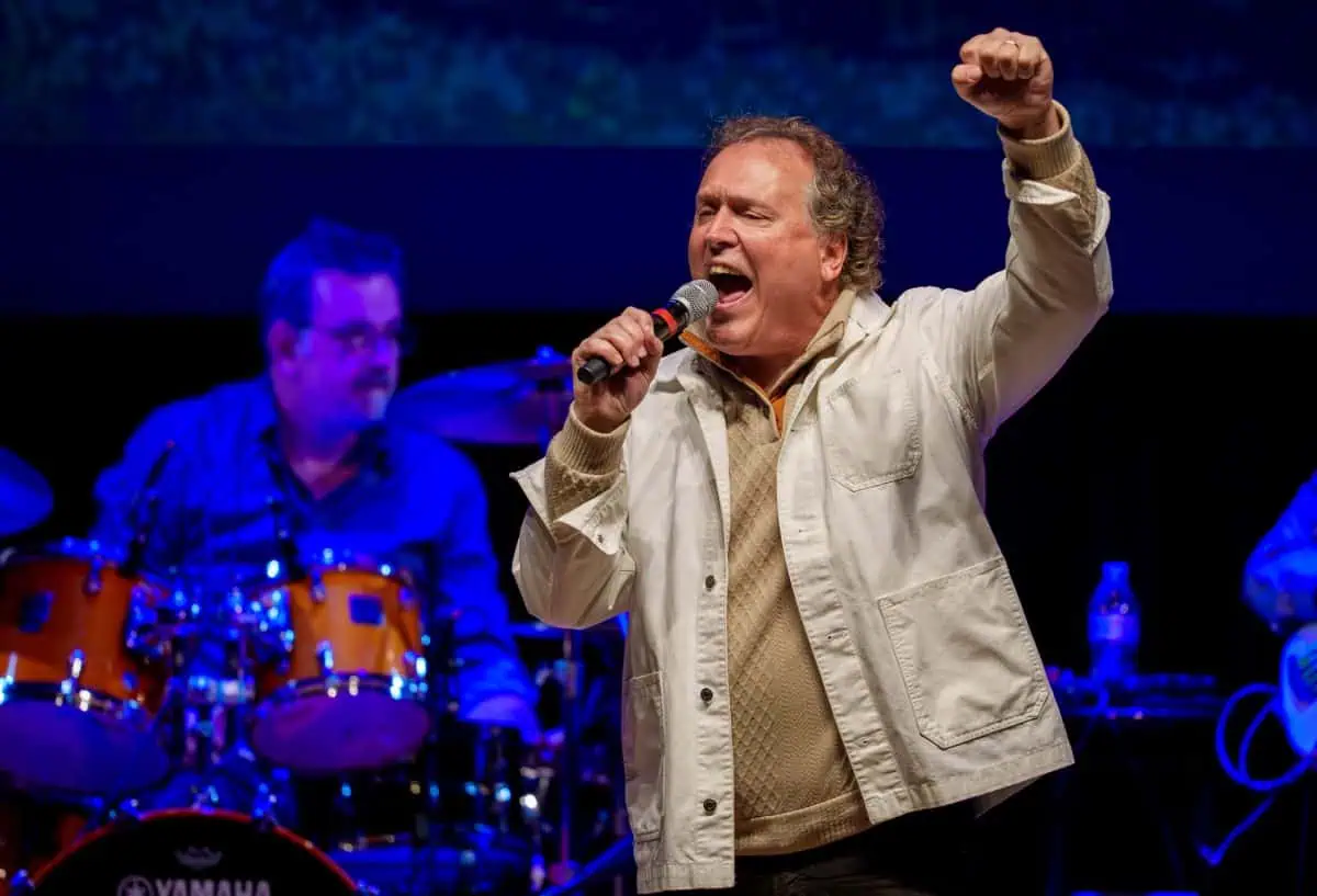 A man in a white jacket sings passionately into a microphone on stage. A drummer plays in the background.