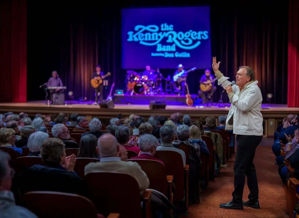 A performer in a white jacket sings to a seated audience in a theater. The stage displays "The Kenny Rogers Band" with musicians playing instruments in the background.