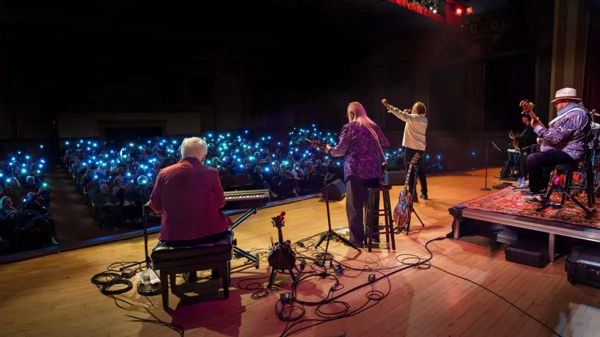 View from behind The Kenny Rogers Band performing on stage with an audience holding up lights. The guitarist and keyboard player are seated, while another musician stands and gestures toward the crowd.