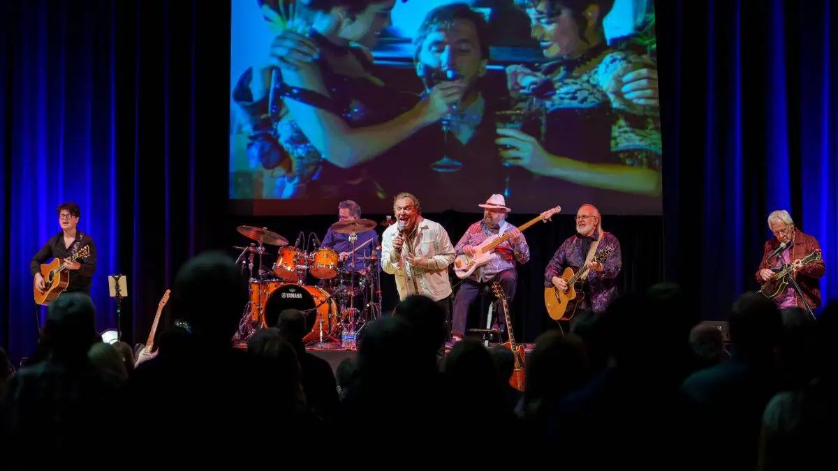 A band performs on stage with a singer and instrumentalists, including guitarists and a drummer. A large screen behind them displays an image with three people in an embrace. Audience members watch.