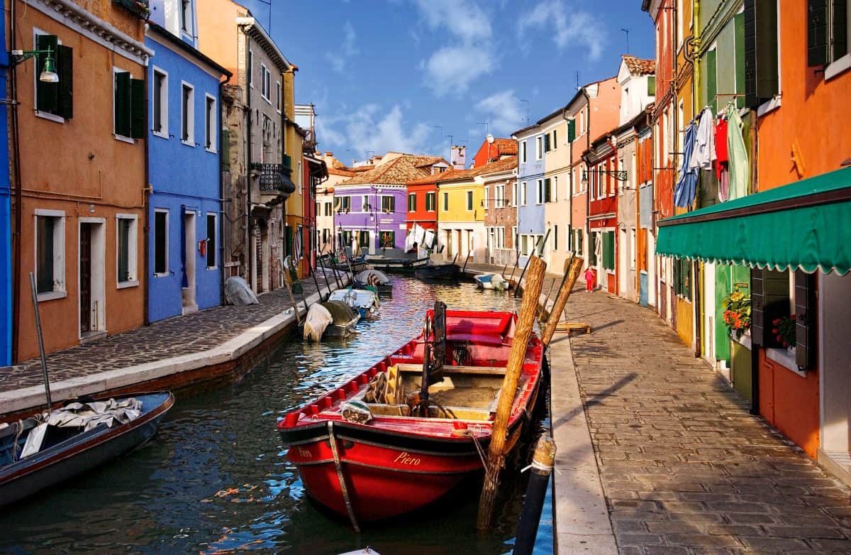 A narrow canal lined with colorful buildings is captured in stunning photography, with boats docked along the water under a partly cloudy sky, highlighting how aging eyes perceive color differently.