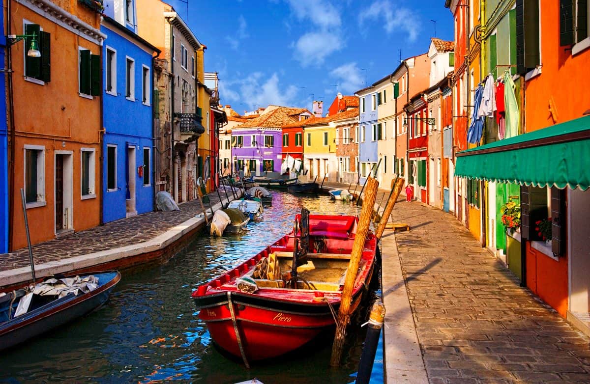 A narrow canal lined with colorful buildings and small boats, including a red boat in the foreground, captures how aging eyes color perception can influence photography under a bright blue sky.