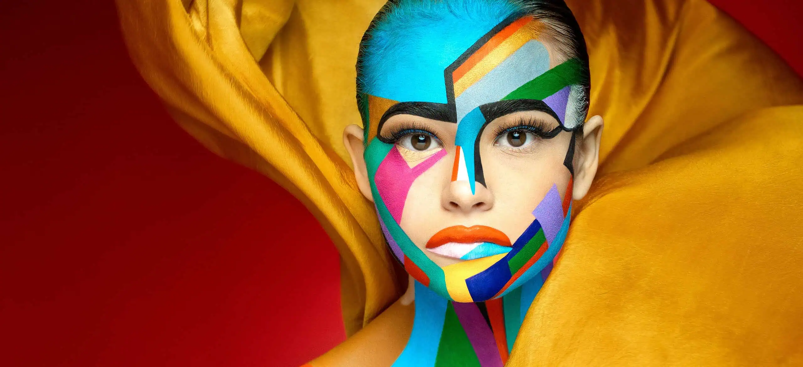 A person with bold, geometric, multicolored face paint poses against a red background, draped in bright yellow fabric.