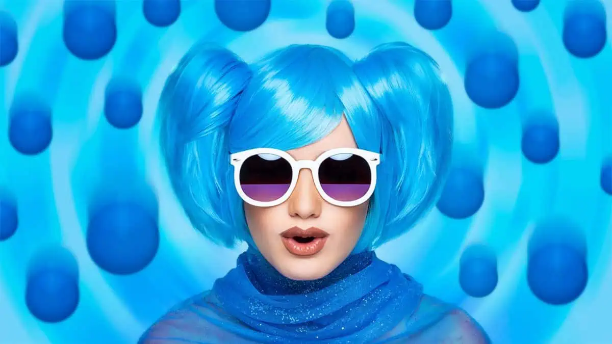 Person with bright blue hair in pigtails, wearing white sunglasses and a blue outfit, poses in front of a blue background with circular patterns and floating blue spheres.