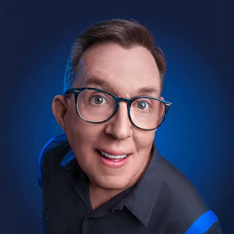 A person with short brown hair, large glasses, and a wide smile is looking up at the camera against a blue gradient background. They are wearing a dark shirt with blue accents.