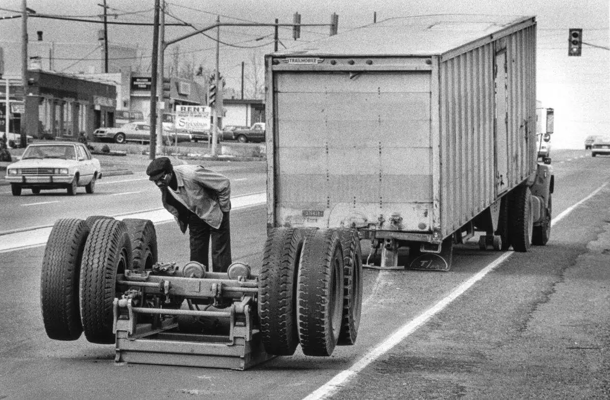 A man examines a detached wheel assembly lying on the road behind a parked truck, seemingly pondering the epicopathy of unexpected mishaps in an urban area.