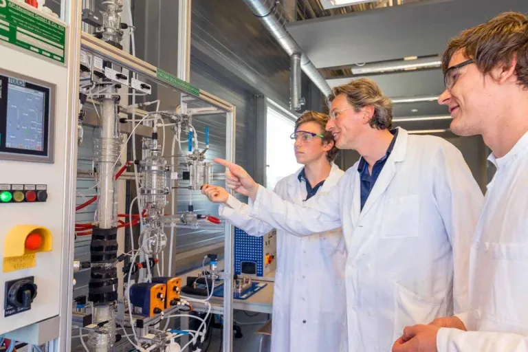 Three people in white lab coats and safety glasses examine and discuss mixing equipment in a laboratory, with one person pointing at a glass apparatus connected to various tubes and wires.