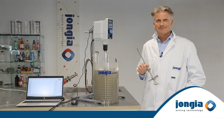 A man in a white lab coat stands beside a mixing machine with a clear tank, holding a metal impeller. A laptop displaying a mixing graph sits on the table, while shelves with bottles and a Jongia banner are in the background.