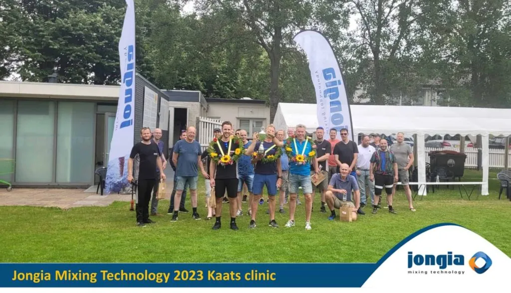 A group of people stand outdoors on grass, some holding sunflowers and medals, posing for a photo in front of banners and tents at the Jongia Mixing Technology 2023 Kaats clinic, celebrating teamwork and mixing tradition with innovation.