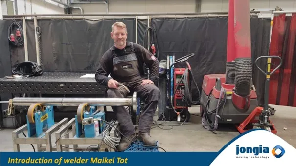A smiling welder in work overalls sits on a metal pipe, surrounded by welding and mixing equipment. A blue banner reads, Introduction of welder Maikel Tot, with the Jongia Mixing Technology logo in the corner.