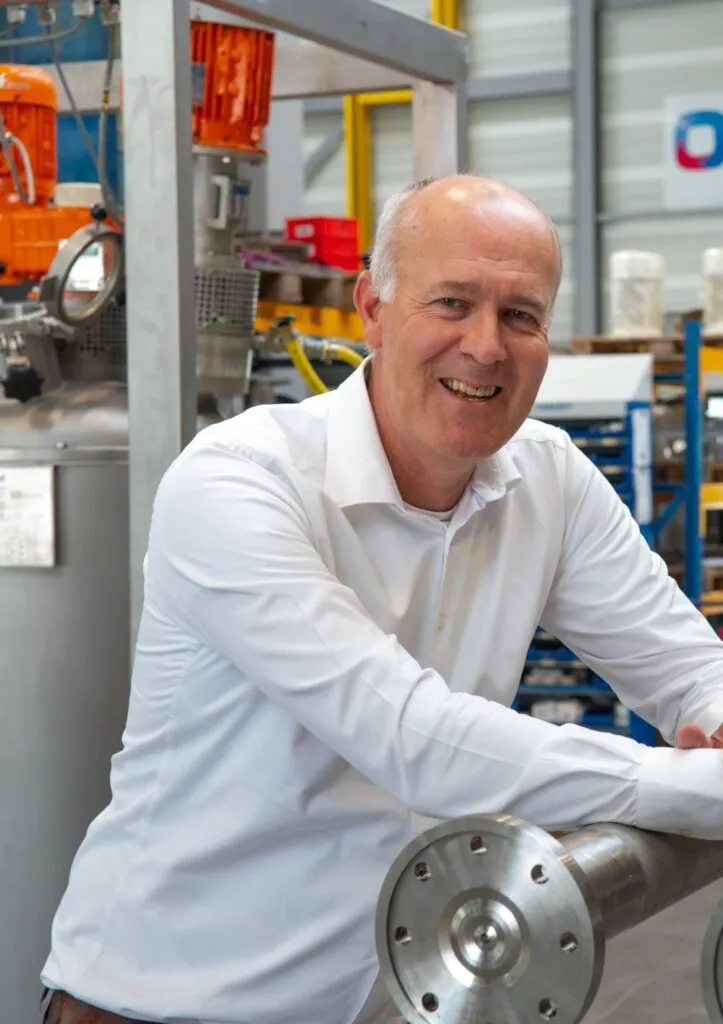 A smiling middle-aged man in a white shirt leans on a large metal mixing cylinder in an industrial workshop with machinery and shelves in the background.