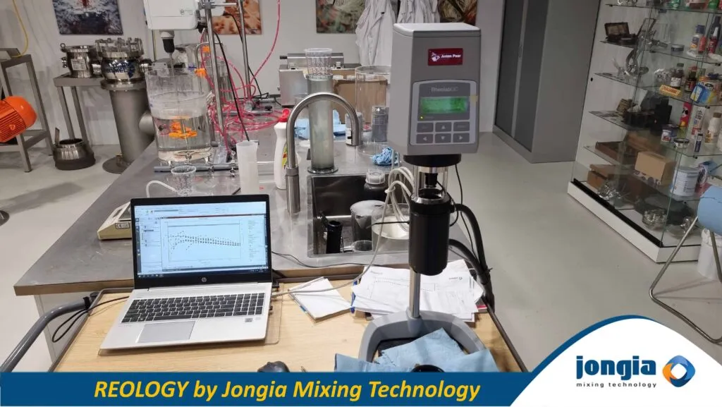 A laboratory workspace with scientific equipment, including a viscometer, mixing apparatus, laptop displaying a graph, glassware, and cables. The image features the Jongia Mixing Technology logo and the text REOLOGY by Jongia Mixing Technology at the bottom.