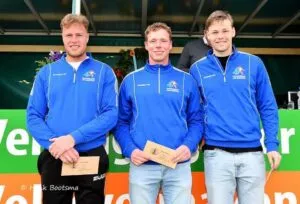 Three young men stand side by side, smiling and wearing matching blue athletic jackets. Mixing excitement with pride, they each hold an envelope and pose in front of a green and orange sign, with flowers visible in the background.
