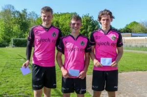 Three young men wearing matching bright pink sports jerseys and black shorts stand on a grassy field, mixing fun with team spirit as they each hold a white envelope and smile at the camera. Trees and a goalpost are in the background.