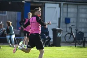 A young man in a pink sports jersey and black shorts is playing soccer outdoors, mixing energy and focus as he prepares to kick the ball. People and a bicycle are visible in the background near a building.