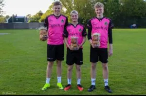 Three young athletes in matching pink and black sports uniforms stand on a grassy field, each holding a wrapped loaf of bread as a prize for mixing skill and teamwork, smiling at the camera. Trees and buildings are visible in the background.
