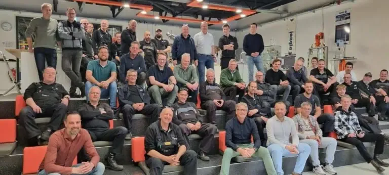 A large group of people, mostly men, are seated and standing on tiered steps in a modern, industrial indoor setting. They are casually dressed, some in uniforms. The background shows technical equipment and office elements. All are looking at the camera and smiling.