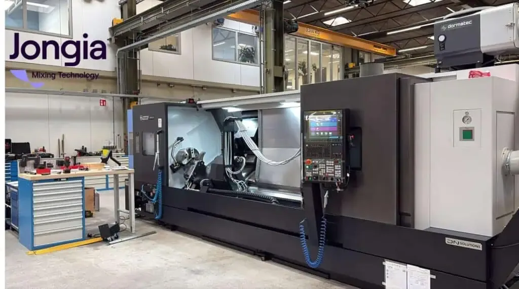 A large CNC machine stands in an industrial workshop with tools and equipment nearby. The machine has a digital control panel, and the Jongia Mixing Technology logo is visible on the wall. Overhead cranes and shelving are in the background.