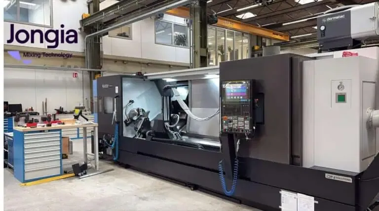A large CNC machine stands in an industrial workshop with tools and equipment nearby. The machine has a digital control panel, and the Jongia Mixing Technology logo is visible on the wall. Overhead cranes and shelving are in the background.