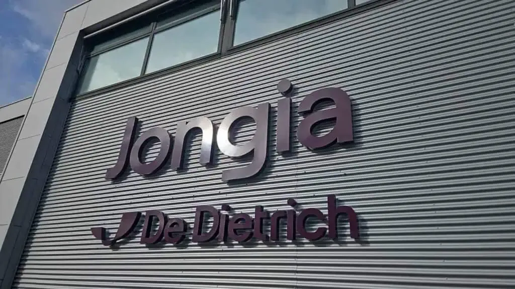 A corrugated metal building facade with large, dark purple 3D letters spelling “Jongia” above a smaller “De Dietrich” logo and text. The building has large windows above the sign and a blue sky is visible in the upper left corner.