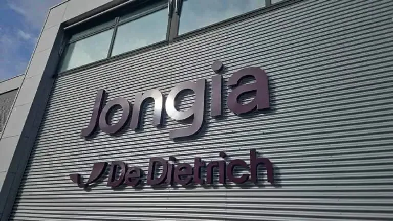 A corrugated metal building facade with large, dark purple 3D letters spelling “Jongia” above a smaller “De Dietrich” logo and text. The building has large windows above the sign and a blue sky is visible in the upper left corner.