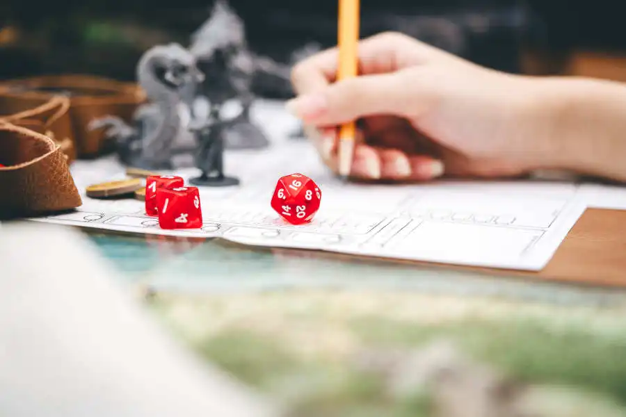 Eine Fotografie einer Hand die einen Stift hält und von Würfeln, Figuren und Pen and Paper Notizen umgeben ist.