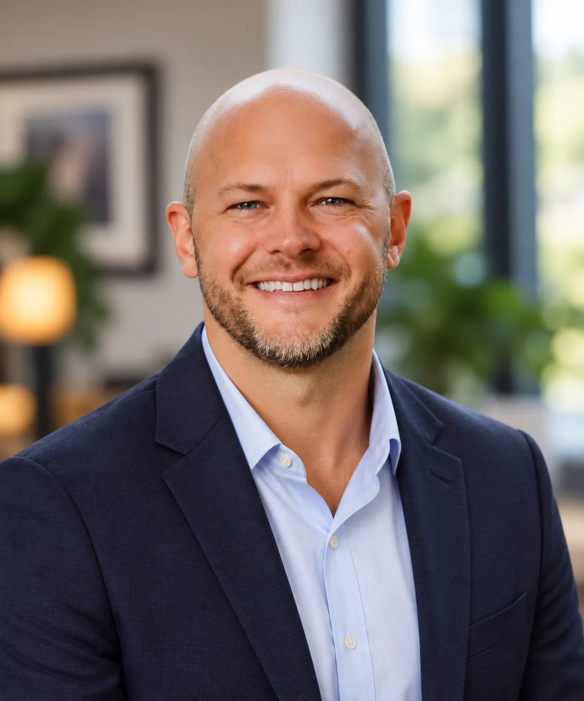 A smiling bald man with a short beard, wearing a navy blazer and light blue shirt, sits in a modern office with blurred plants and windows in the background, embodying the professional confidence of a Pittsburgh SEO expert.