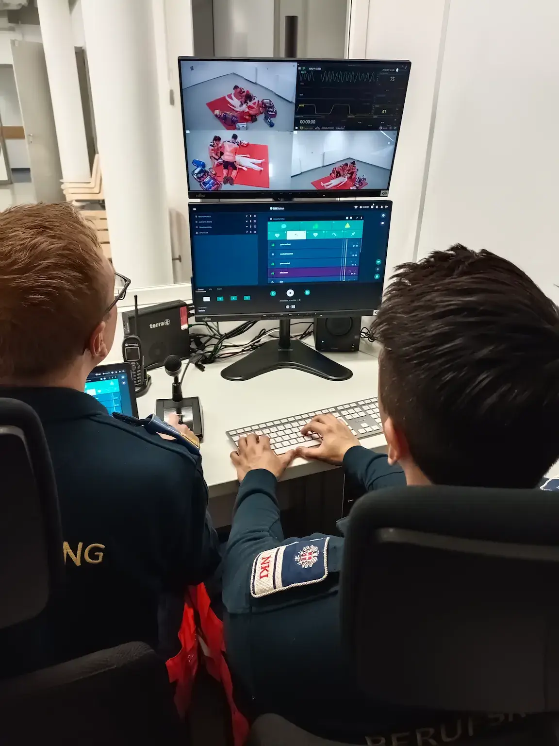 Emergency training personnel at workstation monitoring medical simulation feeds and vital signs on dual screens.