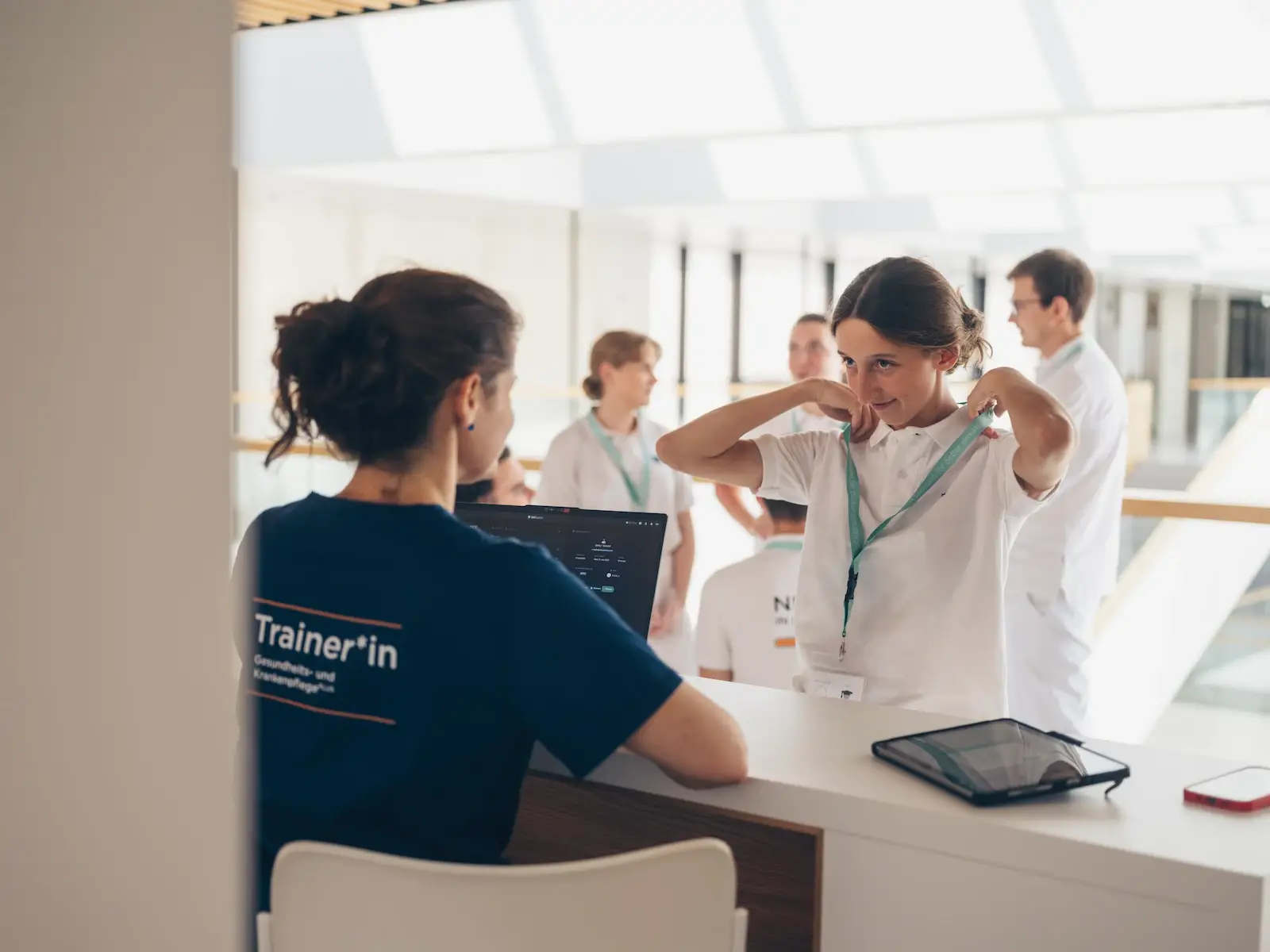 Trainer in nursing education leading healthcare workshop with laptop and tablet, professionals in white uniforms engaged in medical training discussion.