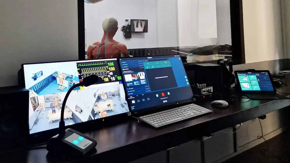 A control room with multiple monitors displays medical data and video feeds. Beyond a window, a medical mannequin is visible in a simulated clinical space, powered by SIMStation Integrated Systems’ highly configurable AV infrastructure for training environments.