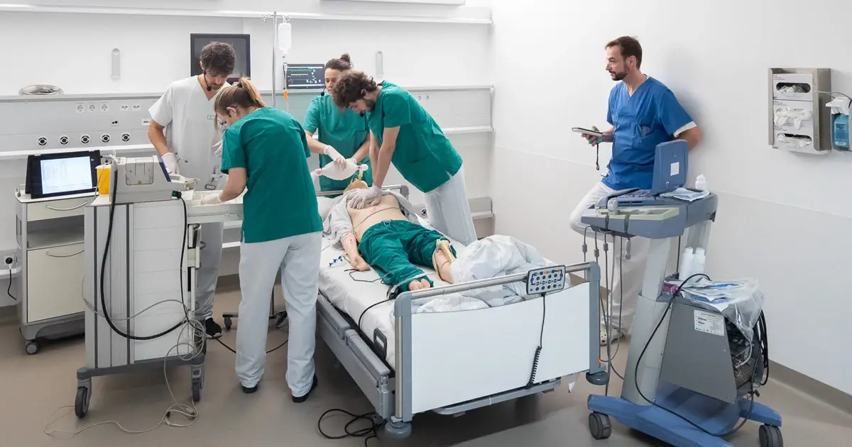 Healthcare professionals practicing clinical procedure on mannequin with tablet monitoring in hospital simulation room.