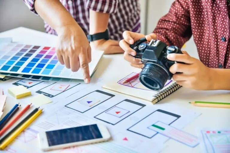 Twee mensen werken aan een bureau bedekt met kleurstalen, een camera, een smartphone en geprinte wireframe-ontwerpen. De ene persoon wijst naar de papieren terwijl de andere de camera vasthoudt, wat een creatieve planningssessie suggereert.