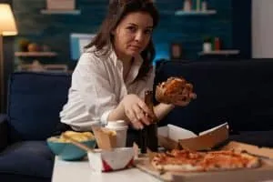 Woman holding beer bottle eating tasty takeaway hamburger while relaxing on couch