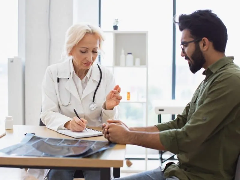 Bearded gentleman describing symptoms of illness to senior female doctor and gesturing hands. Mature elder physician giving recommendations and using notebook for writing prescription.