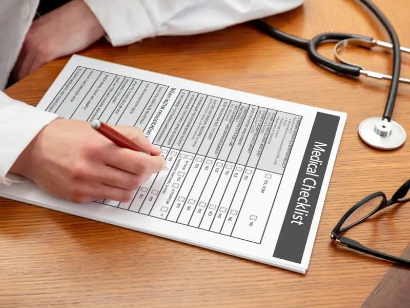 Medical checklist. Woman doctor holding a pen, close up view, wooden office table background. Female hand filling in the document, copy space