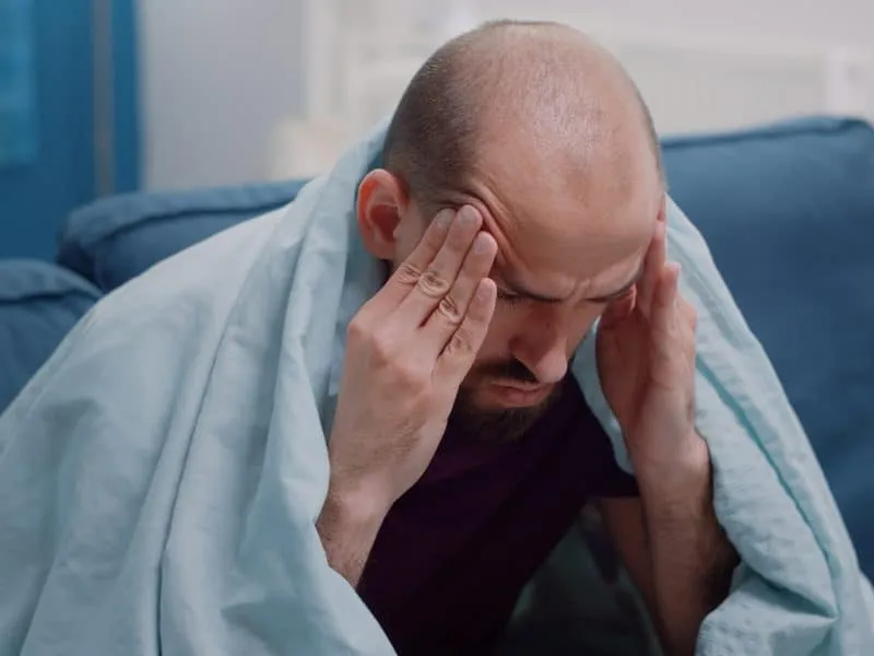 Close up of ill man holding hands on head against migraine. Sick person rubbing temples to cure headache and virus symptoms. Adult having medicaments, bottles of pills, tea and tissues