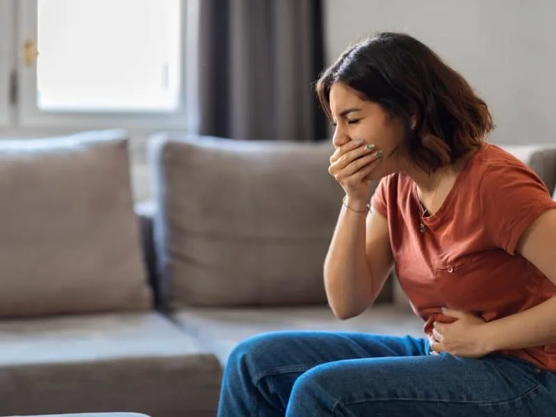 Portrait Of Young Arab Woman Suffering From Nausea At Home, Millennial Middle Eastern Female Having Morning Sickness, Ill Lady Sitting On Couch In Living Room, Covering Mouth And Touching Belly