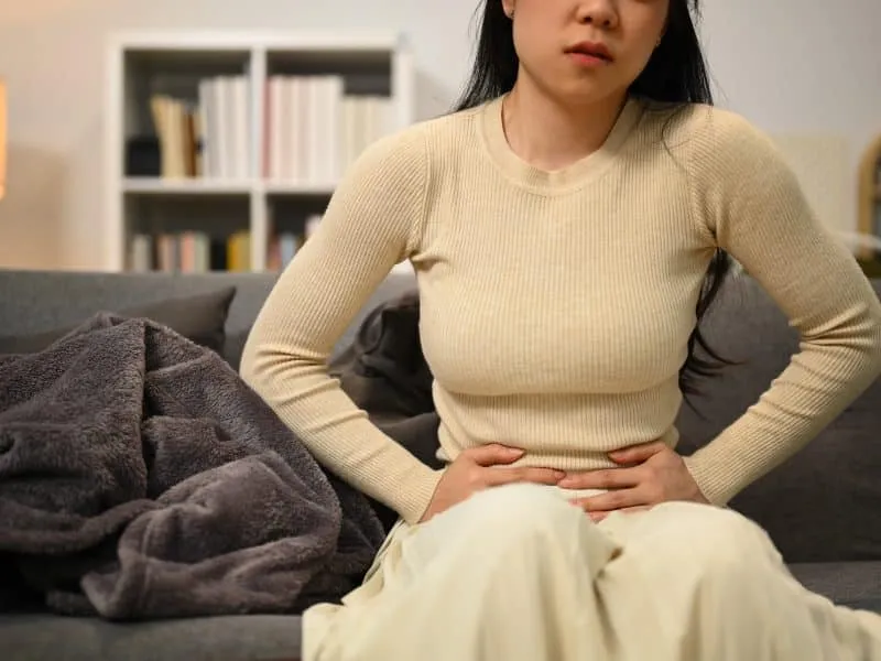 Sick young woman suffering from abdominal pain while sitting on sofa at home. Medical, gynecological problems, health care concept.