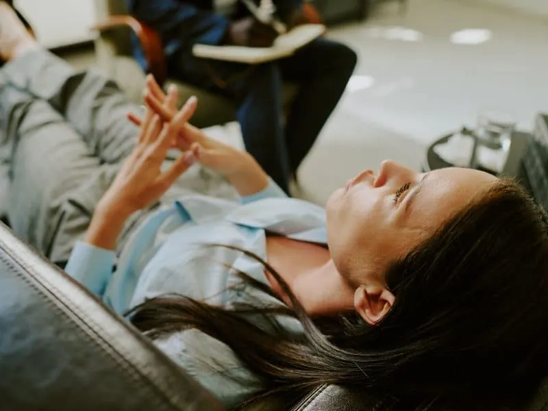 Person lying on couch in therapist's office, engaging in conversation with a professional. Therapist sits across from patient, taking notes