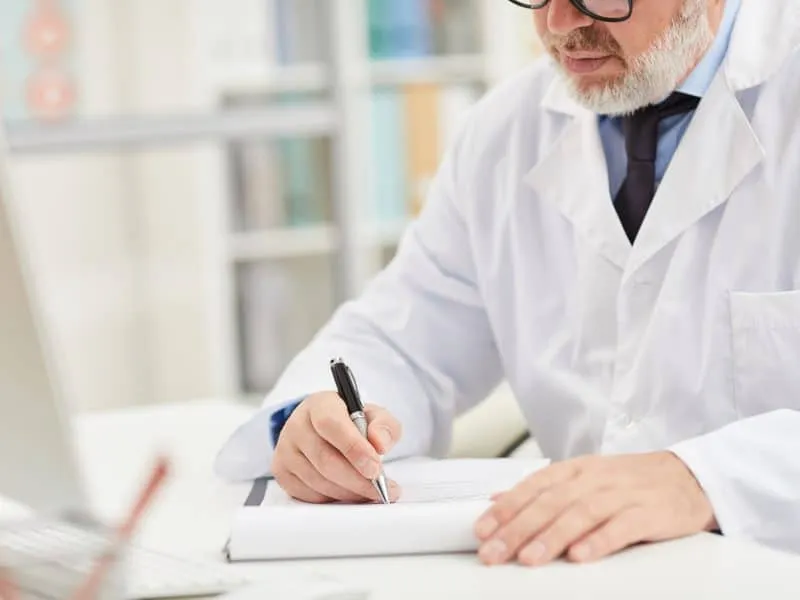 Close-up of male doctor in white coat sitting at the table and doing paper work at office
