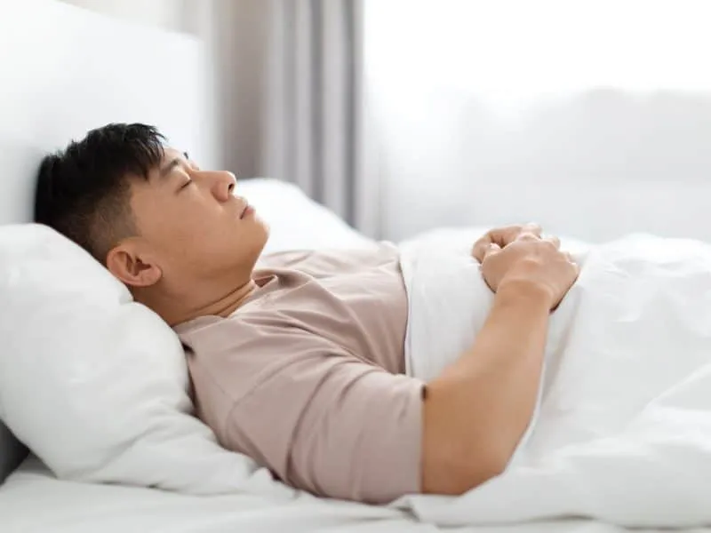 Single handsome middle aged chinese man wearing pajamas peacefully sleeping alone in bed at home, enjoying his comfortable bed, orthopedic mattress and pillow, side view. Healthy sleep concept