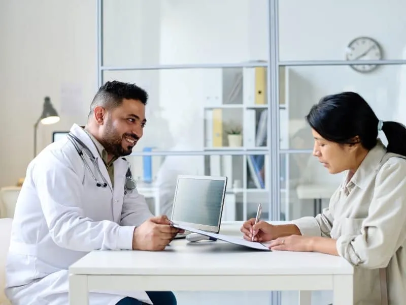 Young woman signing agreement for treatment while sitting at table with doctor in hospital