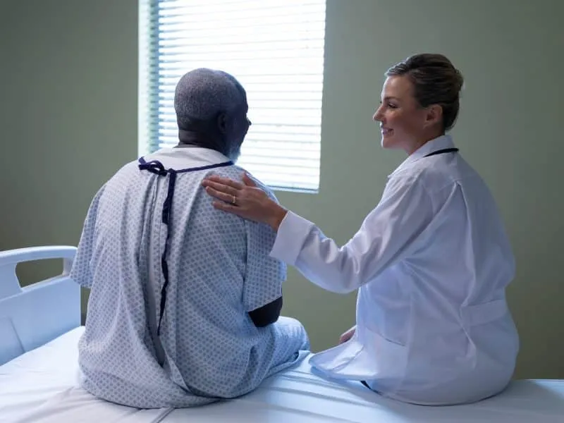 Diverse female doctor and male patient sitting on hospital bed smiling to each other. medicine, health and healthcare services.